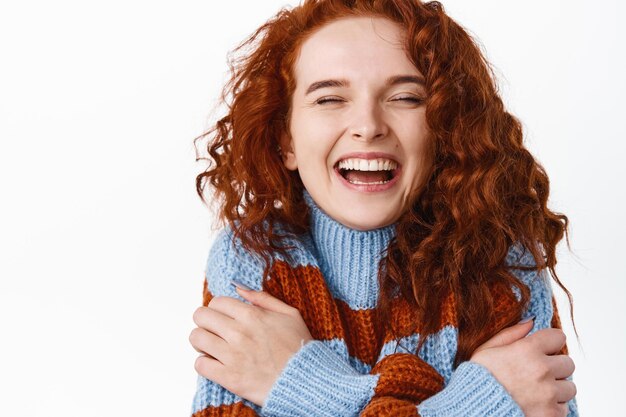 Cerrar retrato de mujer pelirroja hermosa feliz con cabello natural rizado, piel suave y pálida, riendo sinceramente con los ojos cerrados y abrazándose a sí misma en blanco