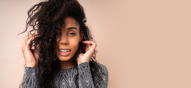 Cerrar retrato de mujer riendo con piel morena y peinado africano en vestido de fiesta brillante posando sobre pared beige.