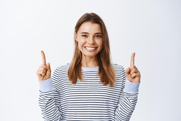 Cerrar retrato de mujer rubia sonriente apuntando con el dedo hacia arriba, mostrando publicidad, dando recomendaciones, demostrando oferta promocional, blanco