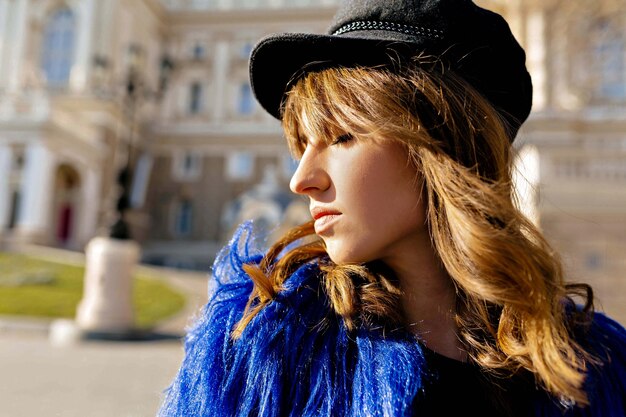 Cerrar el retrato de perfil fuera de la tierna mujer de gorra negra y abrigo azul disfrutando del sol con una sonrisa