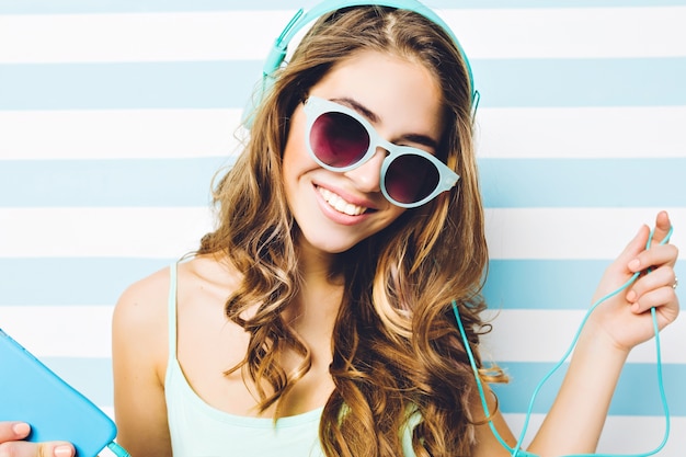 Cerrar retrato de verano con estilo joven atractiva mujer con pelo largo y rizado en gafas de sol azules escuchando música a través de auriculares en la pared azul blanca con rayas. Sonriendo, felicidad.