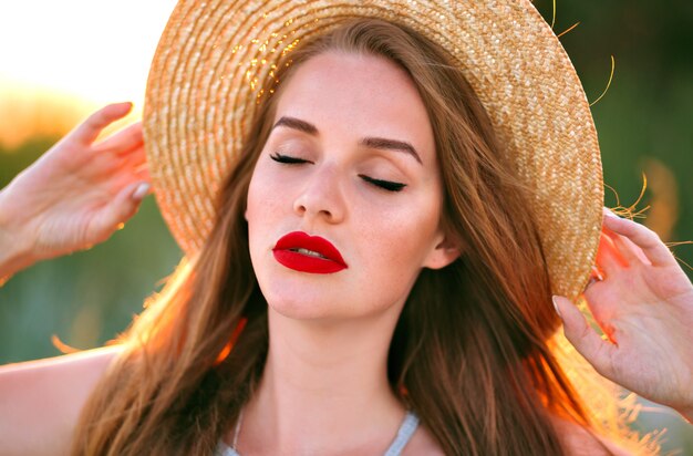 Cerrar tierno retrato de mujer sensual belleza posando en el campo, estilo vintage, con sombrero de paja de moda, maquillaje de belleza natural, rostro pecoso y labios carnosos rojos.