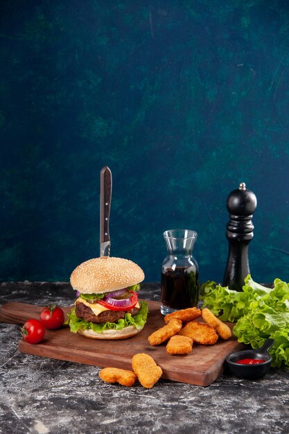 Cerrar vista de cuchillo en sabroso sándwich de carne y nuggets de pollo tomates con tallo pimienta en tablero de madera salsa ketchup sobre superficie azul oscuro