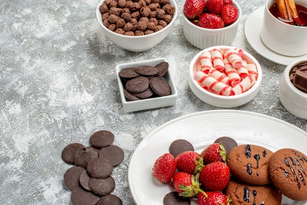 Cerrar vista inferior galletas fresas y bombones redondos en la placa ovalada cuencos con dulces fresas chocolates cereales y té de canela en la mesa gris-blanca