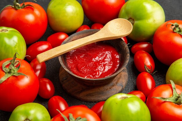 Cerrar vista inferior tomates rojos y verdes cereza alrededor de un recipiente con salsa de tomate y una cuchara de madera sobre fondo oscuro