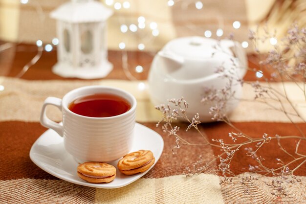 Cerrar vista lateral de una taza de té con galletas té negro en la taza con galletas junto a la tetera y las ramas de los árboles
