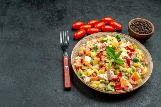 Cerrar vista lateral del tazón de fuente de ensalada de verduras con tenedor, tomates, pimienta sobre tabla gris oscuro
