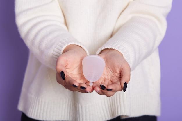 Cerrar vista de mujer sin rostro con camisa blanca con producto de higiene, haciendo la elección de usar copa menstrual o no, posando aislado en púrpura, femenino teniendo período. Concepto de ginecología.