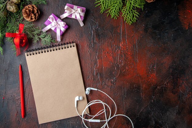 Cerrar vista de ramas de abeto, una taza de té negro, accesorios de decoración, auriculares blancos y regalo junto al cuaderno con bolígrafo sobre fondo oscuro