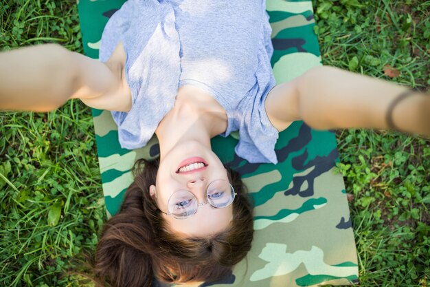 Cerrar vista superior de risa mujer morena en anteojos tumbado en la hierba en el parque y haciendo selfie