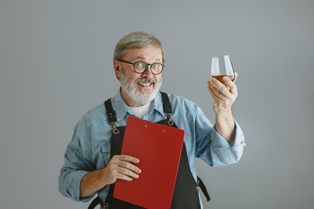 Cervecero hombre mayor seguro con cerveza artesanal en vidrio en barril de madera sobre fondo gris.