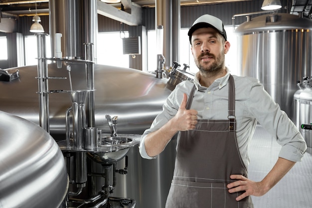 Cervecero profesional en su propia producción artesanal de alcohol. Especialista, hombre vestido con ropa de trabajo posando confiado con el pulgar hacia arriba. Concepto de negocio abierto, producto ecológico, cervecería artesanal, fábrica individual.