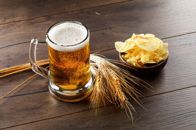 Cerveza con espigas, chips en un vaso en la mesa de madera, vista de ángulo alto.