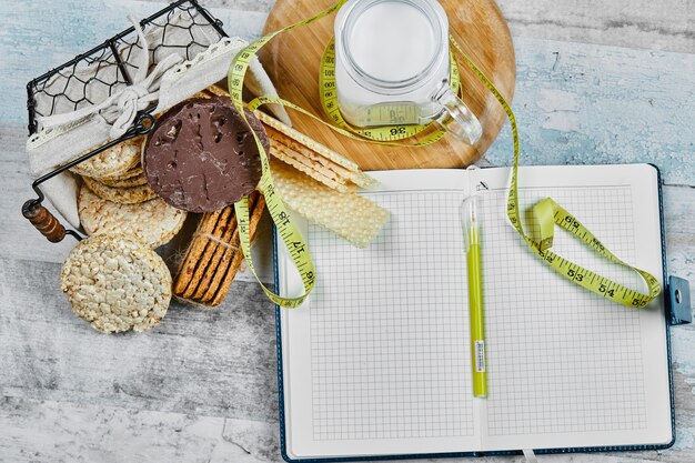 Cesta de galletas y un tarro de leche sobre una mesa de mármol con un cuaderno y un bolígrafo.