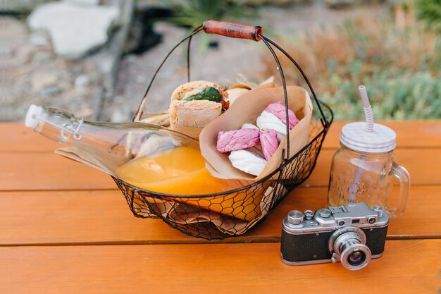 Cesta de hierro con botella de jugo de naranja y bocadillos de pie sobre la mesa de madera. Foto al aire libre de comida para picnic, vaso vacío y cámara.