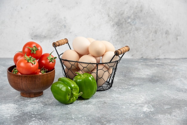 Cesta de mimbre de huevos orgánicos crudos, pimientos y tomates rojos en mármol.