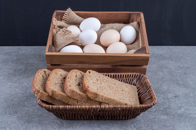 Cesta de mimbre de pan de centeno y caja de madera de huevos crudos en piedra.