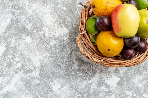 Cesta de mimbre de plástico con vista de la mitad superior con peras de manzana, ciruelas feykhoas y caqui en la parte superior derecha de la mesa gris