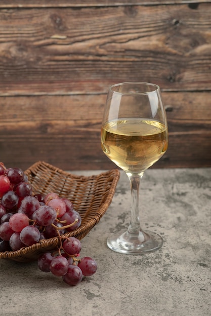 Cesta de mimbre de uvas rojas con copa de vino blanco sobre mesa de mármol.