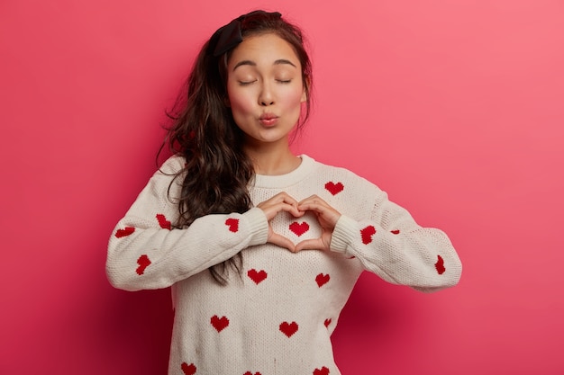Charming woman with folded lips shows heart gesture over chest, expresses heartwarming feelings, sympathy and tender