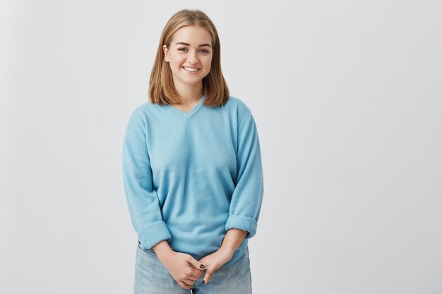 Cheerfulcharming joven vistiendo suéter azul con el pelo liso y rubio sonriendo felizmente mientras recibe algunas noticias positivas. La muchacha bonita se vistió en el azul que miraba con sonrisa alegre.