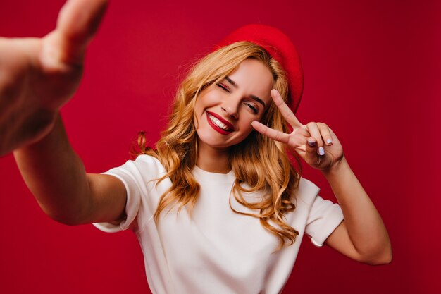 Cheerul mujer guapa haciendo selfie en pared roja. Filmación en interiores de elegante niña francesa despreocupada sonriendo