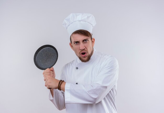 Un chef barbudo enojado hombre vestido con uniforme de cocina blanco y sombrero sosteniendo una sartén como un bate de béisbol en una pared blanca
