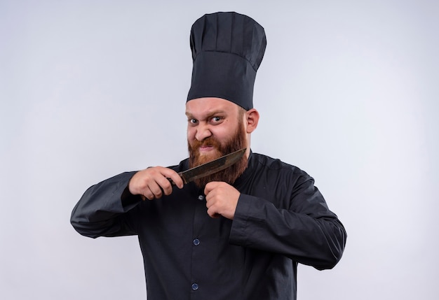 Foto gratuita un chef barbudo enojado con uniforme negro sosteniendo un cuchillo y tratando de cortarse la barba mientras mira a la cámara en una pared blanca