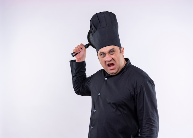 Chef cocinero enojado con uniforme negro y sombrero de cocinero balanceando una sartén con expresión agresiva de pie sobre fondo blanco.