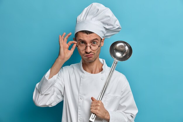 Chef cocinero posa en la cocina comercial, mira seriamente a través de vasos, sostiene un cucharón, prepara comida, plato listo para cocinar, escucha consejos