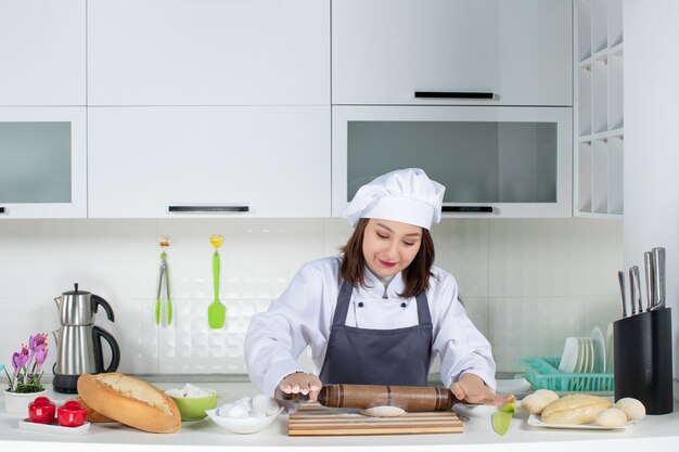Chef de comis femenino concentrado en uniforme de pie detrás de la mesa preparando pasteles en la cocina blanca