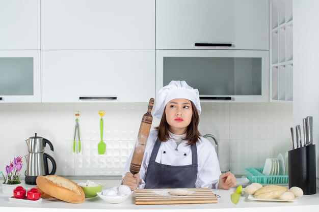 Chef de comis femenino confuso en uniforme de pie detrás de la mesa preparando pasteles en la cocina blanca