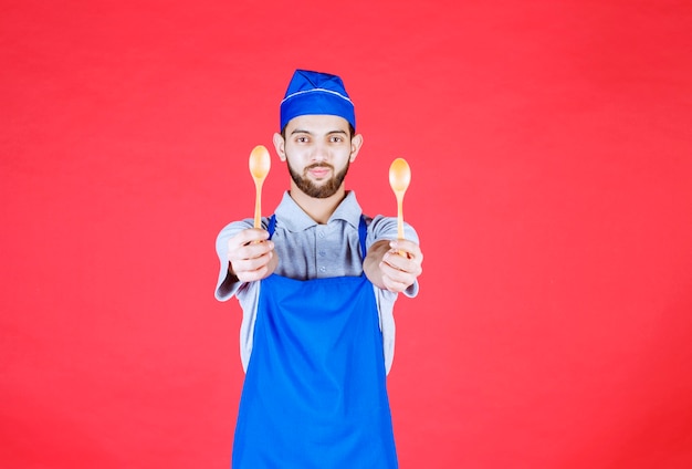 Chef en delantal azul sosteniendo pequeñas cucharas de madera y ofreciéndolas a su colega.