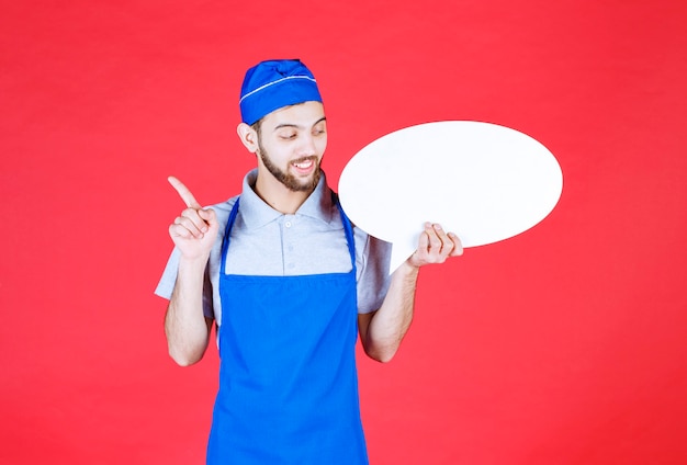 Chef en delantal azul sosteniendo un tablero de información ovale y teniendo una buena idea.