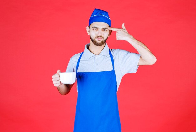 Chef en delantal azul sosteniendo una taza de cerámica blanca y pensando.