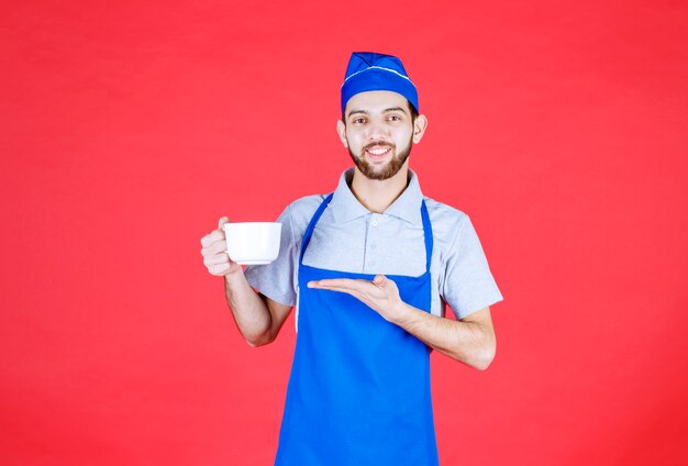 Chef en delantal azul sosteniendo una taza de cerámica blanca.