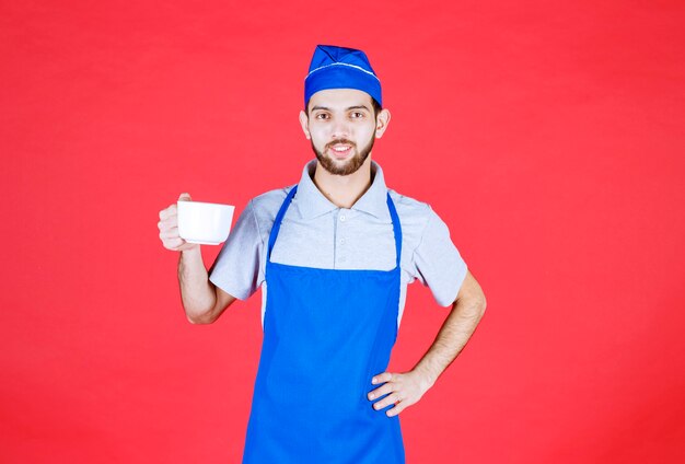 Chef en delantal azul sosteniendo una taza de cerámica blanca.