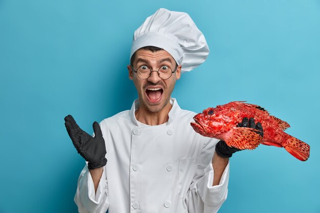 El chef enojado grita en voz alta, mantiene la boca abierta, usa uniforme de cocinero, sostiene un pez grande, da una clase magistral de cocina deliciosa cocina, dice la receta perfecta