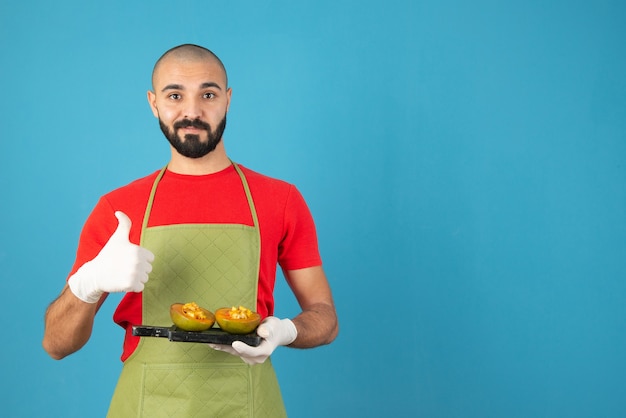 Chef hombre barbudo en delantal y guantes sosteniendo una tabla de madera oscura con pastelería.