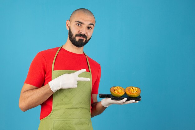 Chef hombre barbudo en delantal y guantes sosteniendo una tabla de madera oscura con pastelería.