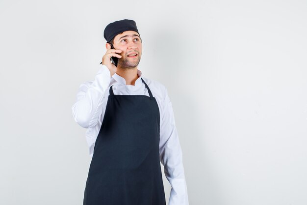 Chef hombre hablando por teléfono inteligente mientras mira hacia arriba en uniforme, delantal, vista frontal.