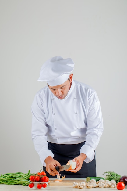 Chef hombre pelando cebolla sobre tabla de madera en uniforme, delantal y sombrero en la cocina