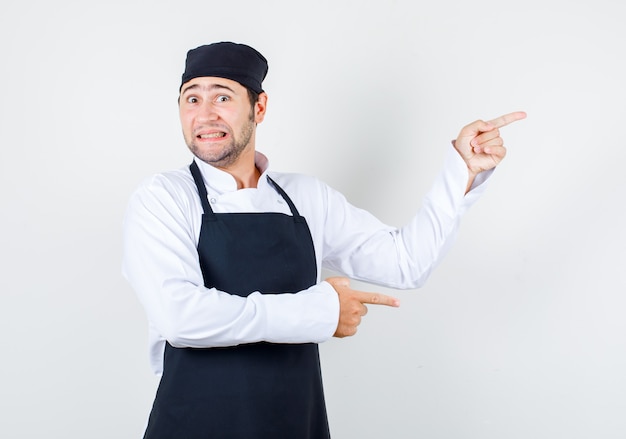 Chef hombre señalando con el dedo a los lados en uniforme, delantal y mirando asustado, vista frontal.
