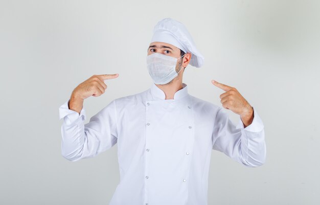 Chef hombre señalando con el dedo a la máscara médica en uniforme blanco y mirando con cuidado