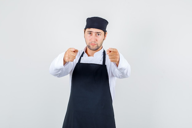 Chef hombre señalando con el dedo en uniforme, delantal y mirando serio, vista frontal.