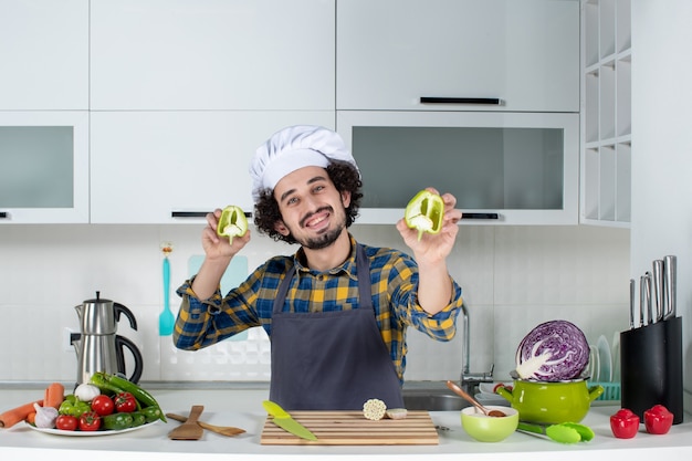Chef hombre sonriente con verduras frescas y cocinar con utensilios de cocina y sosteniendo los pimientos verdes cortados en la cocina blanca