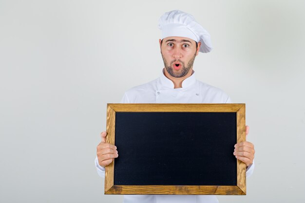 Chef hombre sosteniendo la pizarra en uniforme blanco y mirando conmocionado