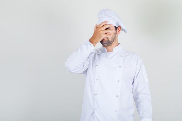 Chef hombre en uniforme blanco mirando a través de los dedos con un ojo y mirando tímido