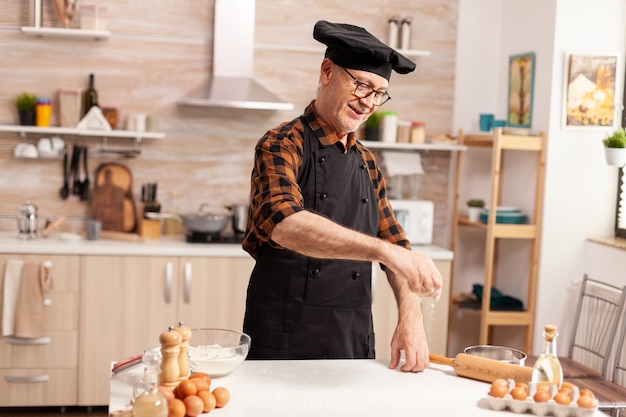 Chef jubilado en la cocina de casa esparciendo harina de trigo sobre la mesa mientras prepara Cook f hecho a mano con bonete y delantal, con uniforme de cocina, tamizado, tamizado, tamizado, ingredientes a mano.