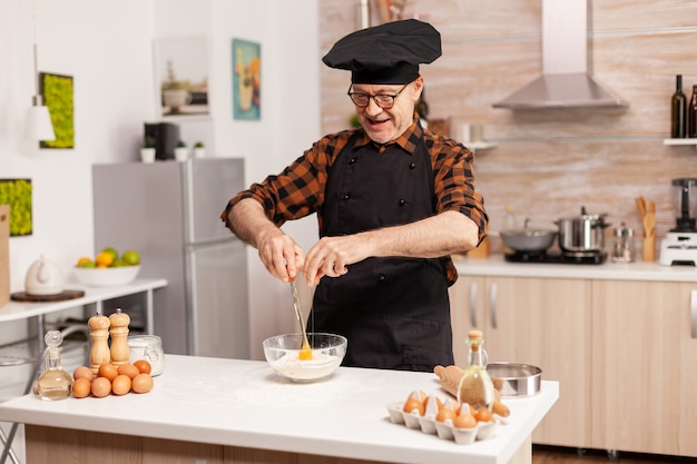 Chef jubilado romper huevos para harina de trigo en la cocina de casa. Anciano pastelero romper el huevo en un tazón de vidrio para la receta de la torta en la cocina, mezclar a mano, amasar.